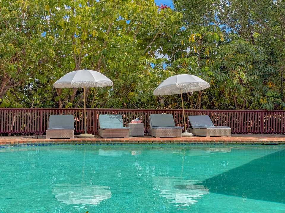 Saltwater pool with four cushioned loungers under white fringe umbrellas, backed by tall trees