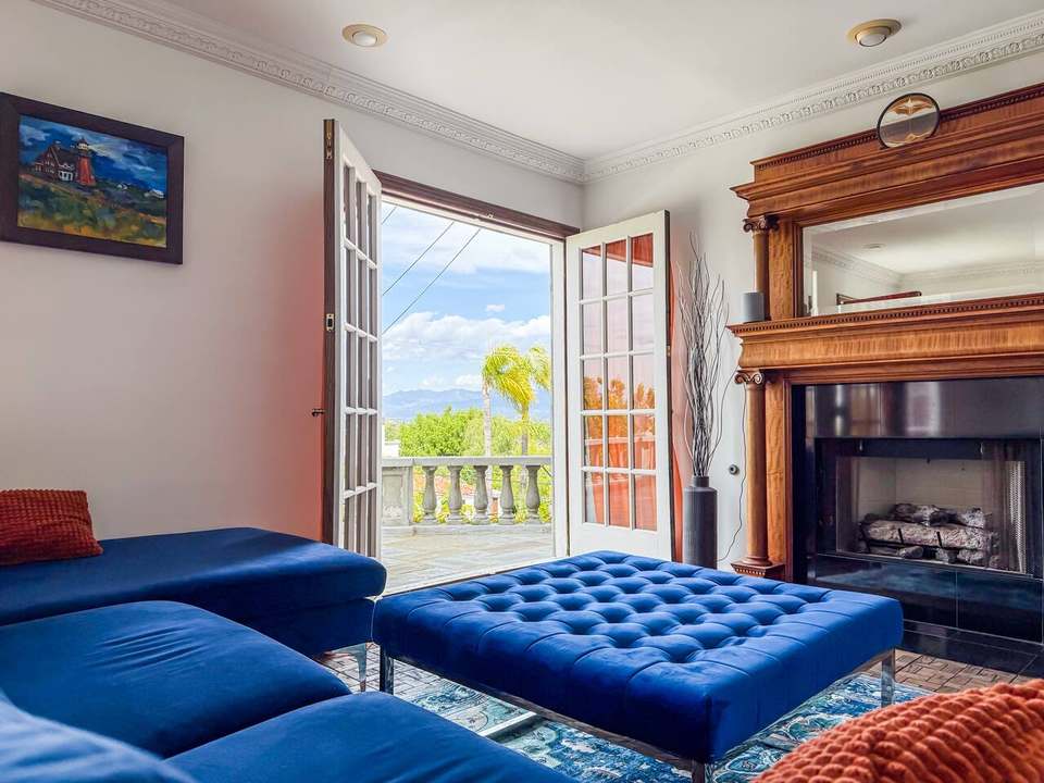 Family room with a blue velvet sectional, tufted ottoman and carved wood fireplace, French doors open to the terrace