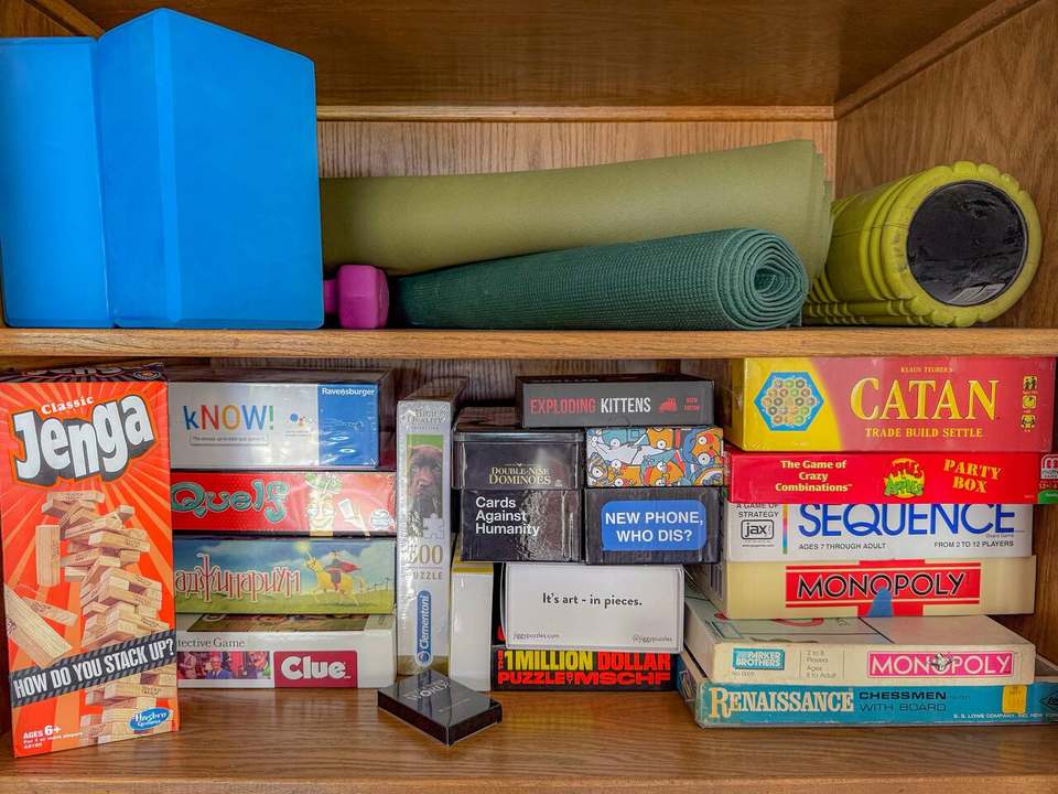 Family-room shelf of board games (Catan, Jenga, Clue and more) with yoga mats and foam rollers